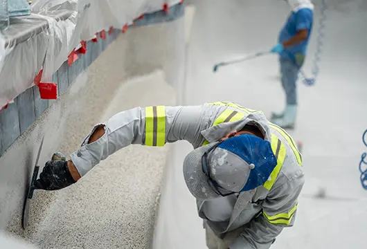 Worker applying textured plaster to the interior wall of an in-ground swimming pool while wearing a high-visibility shirt and protective gear, with another worker spraying material in the background.
