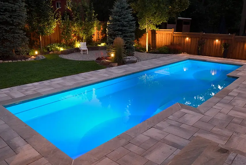 Illuminated rectangular backyard swimming pool at night with blue underwater lighting, surrounded by stone pavers, landscaped garden beds, a wooden fence, and soft outdoor accent lights.