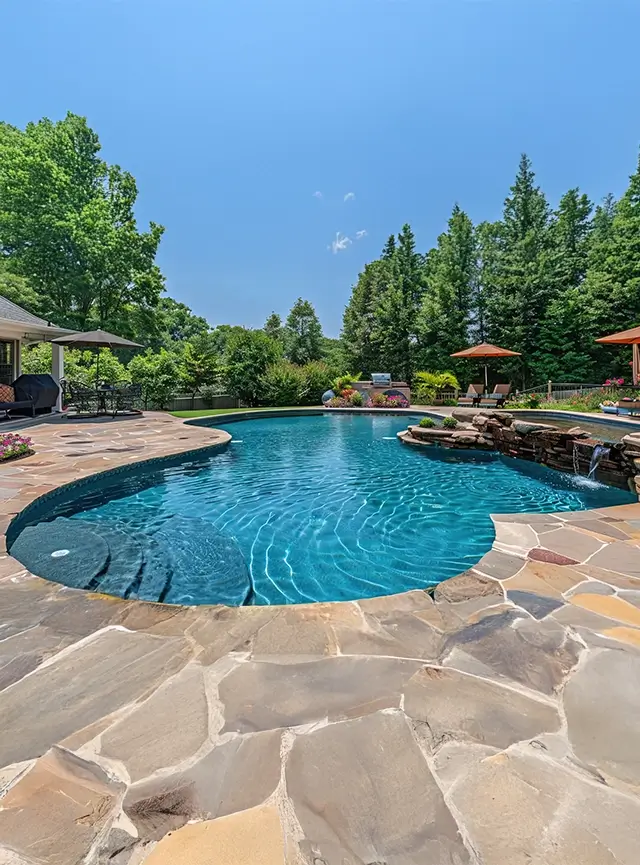 Large backyard swimming pool with natural stone decking and a small rock waterfall feature, surrounded by lush trees, patio seating, and umbrellas under a clear blue sky.